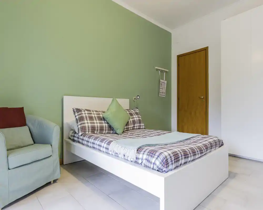 Bright, neatly styled bedroom with a white platform bed, patterned bedding and a mint-green accent wall; includes a comfortable armchair and built-in wardrobe.
