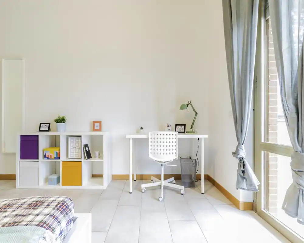 Bright, minimal bedroom corner with a visible bed corner, a white desk and shelving. Large window with tied-back curtains provides strong natural light and a neat, modern feel.