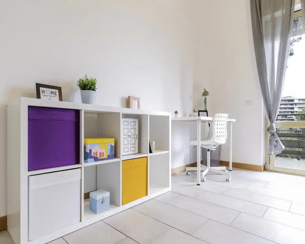 Bright, minimal bedroom corner with open shelving and a small desk by a window — clean lines, colorful storage boxes and natural light ideal for a workspace within the bedroom.