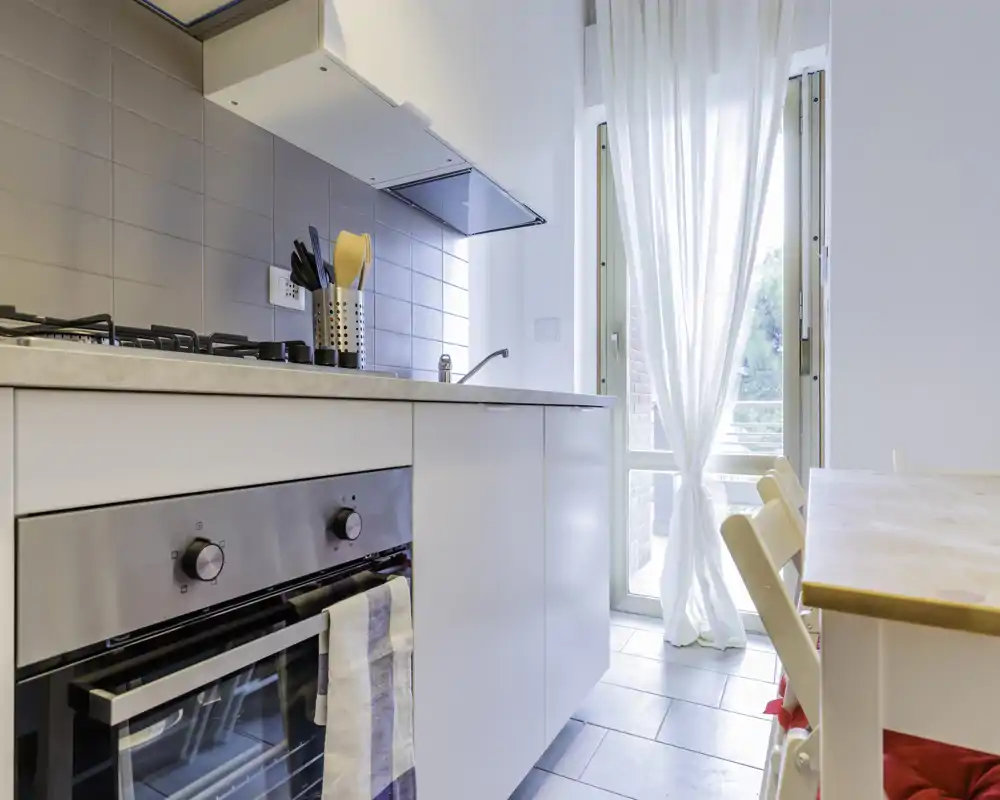 Bright modern kitchen with a built-in oven, gas hob and light-coloured cabinetry; natural light floods the space through a glass door with sheer curtains, next to a small dining area.