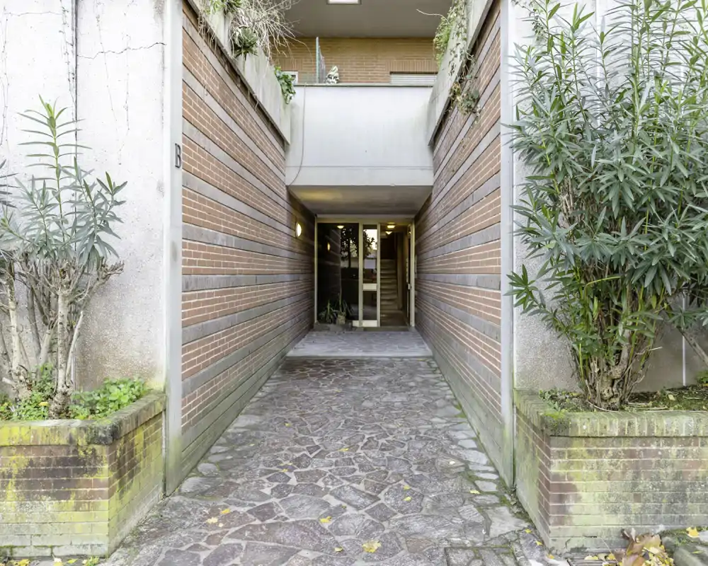 Covered entryway with a paved path leading to a building entrance, flanked by brick planters and greenery; well-lit and symmetrically framed.