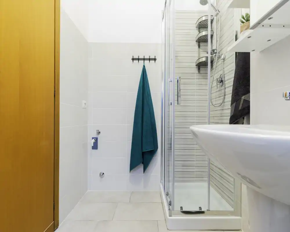 Bright, modern bathroom with a glass-enclosed shower, white sink and teal towel on the wall hook. Clean tiles and contemporary fixtures create a fresh, functional space.