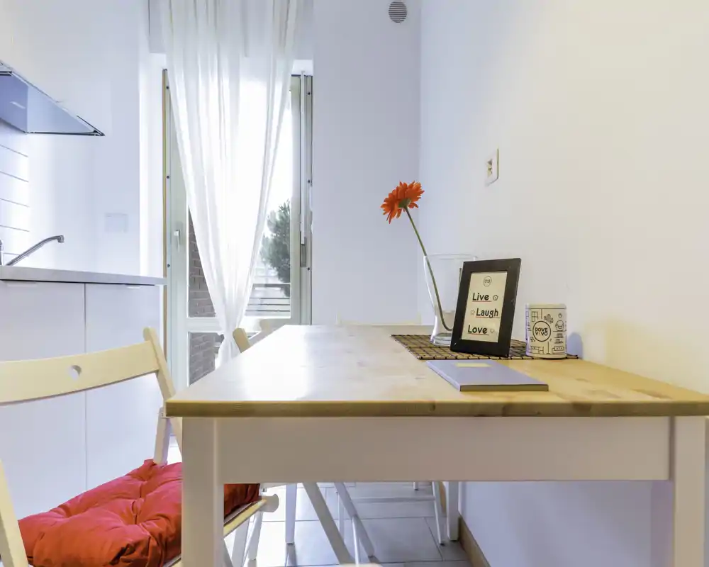Bright, minimalist dining corner with a wooden table, two chairs (one with a red cushion), a decorative vase and framed sign—ideal for casual meals.