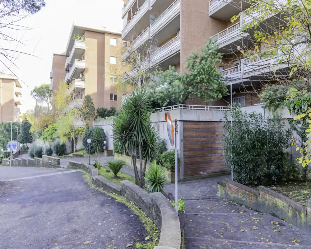 Exterior view of a residential complex entrance with paved walkways, low walls, greenery and balconies; bright daylight and clear architectural details.