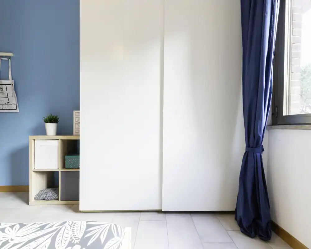 Bright, minimalist bedroom corner with a visible bed corner, a white wardrobe, blue accent wall and curtain — clean modern styling ideal for listings.