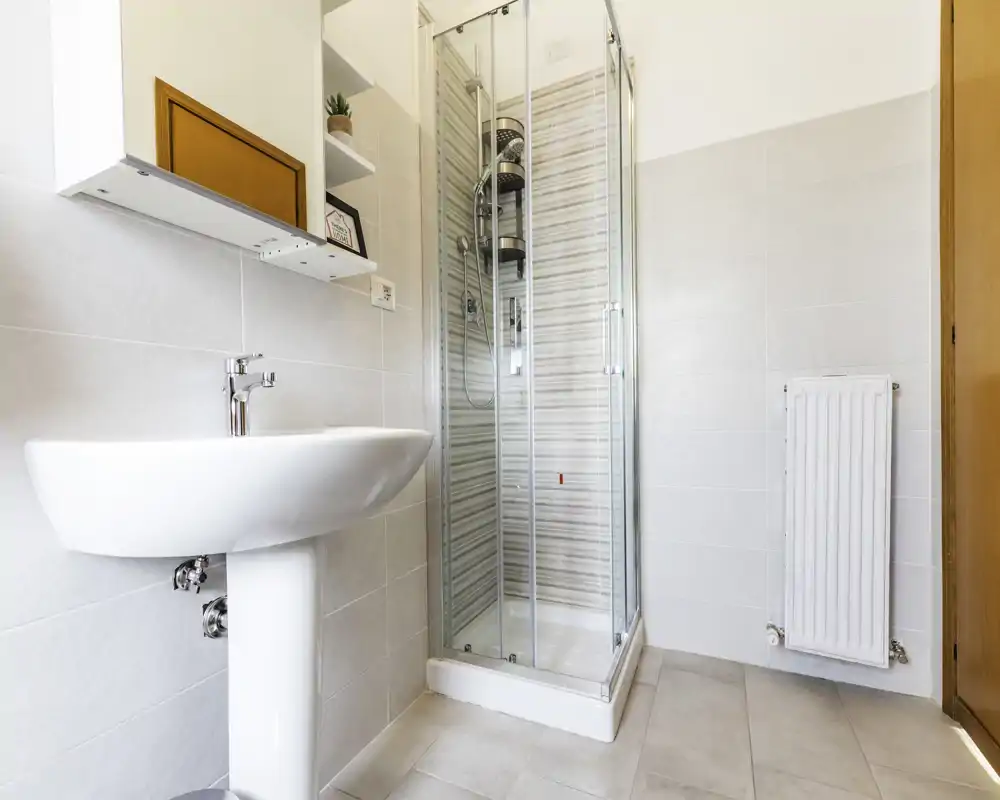Bright, modern bathroom with a pedestal sink and a glass-enclosed corner shower. Neutral tiles and a wall radiator complete the clean, functional space.
