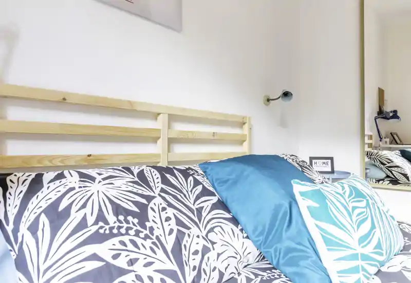 Bright, stylish bedroom corner with a wooden slatted headboard and patterned bedding accented by teal cushions. The photo highlights bed textiles and a cozy, modern bedside styling.