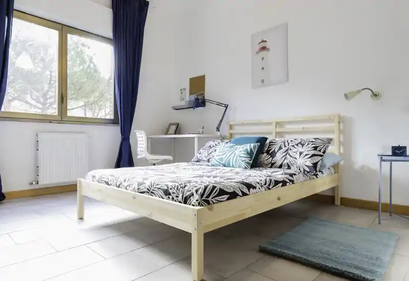 Bright, airy bedroom with a natural wood bed frame, patterned bedding and decorative cushions. A large window and neat desk create a comfortable workspace and pleasant natural light.