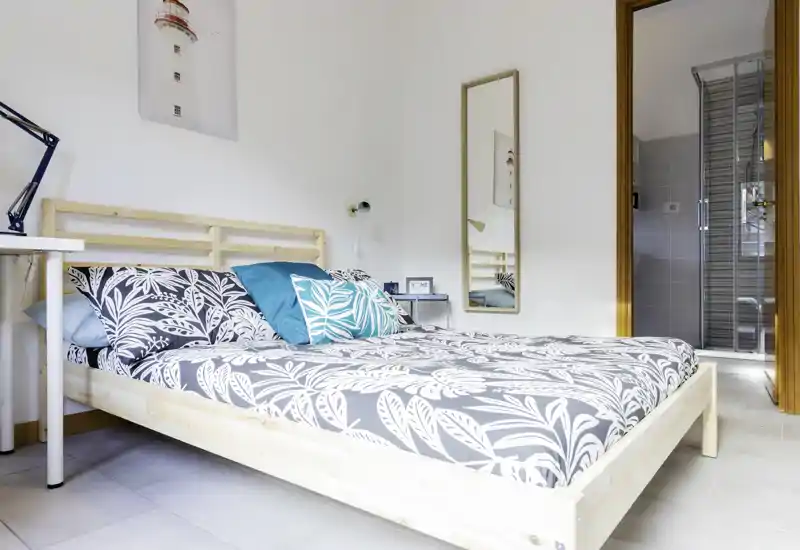 Bright, modern bedroom with a full wooden bed frame, patterned bedding and decorative cushions; clean lines and a small en-suite visible through the door.