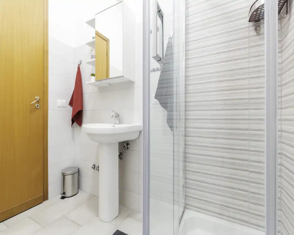 Bagno moderno e luminoso con lavabo a colonna, specchiera con mobiletto e box doccia in vetro con piastrelle strutturate.