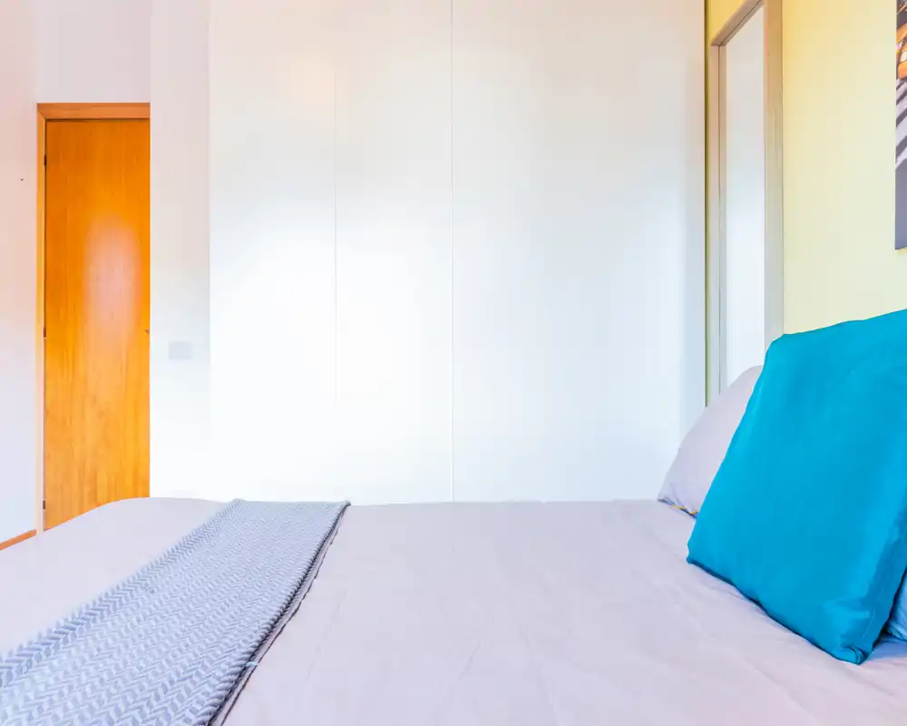 Camera da letto luminosa e minimal con letto ben rifatto, cuscini colorati e un armadio bianco sullo sfondo — perfetta per un annuncio incentrato sull’area notte.