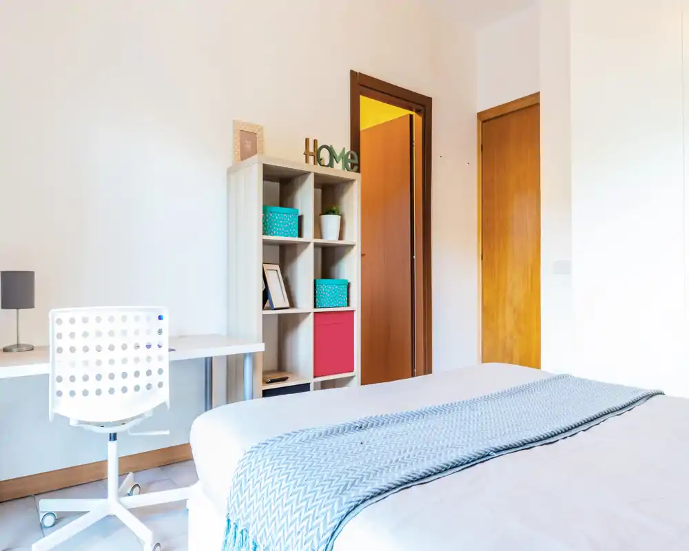 Camera da letto luminosa e minimal con letto ben rifatto, tessuti chiari e una scrivania con libreria: ideale per una persona o uno studente.