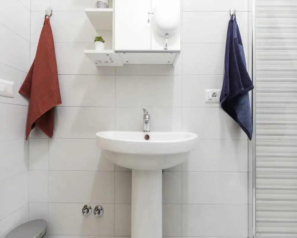 Bagno moderno e luminoso con lavabo a colonna, pensile e asciugamani; piastrelle bianche che conferiscono un aspetto pulito e minimalista.