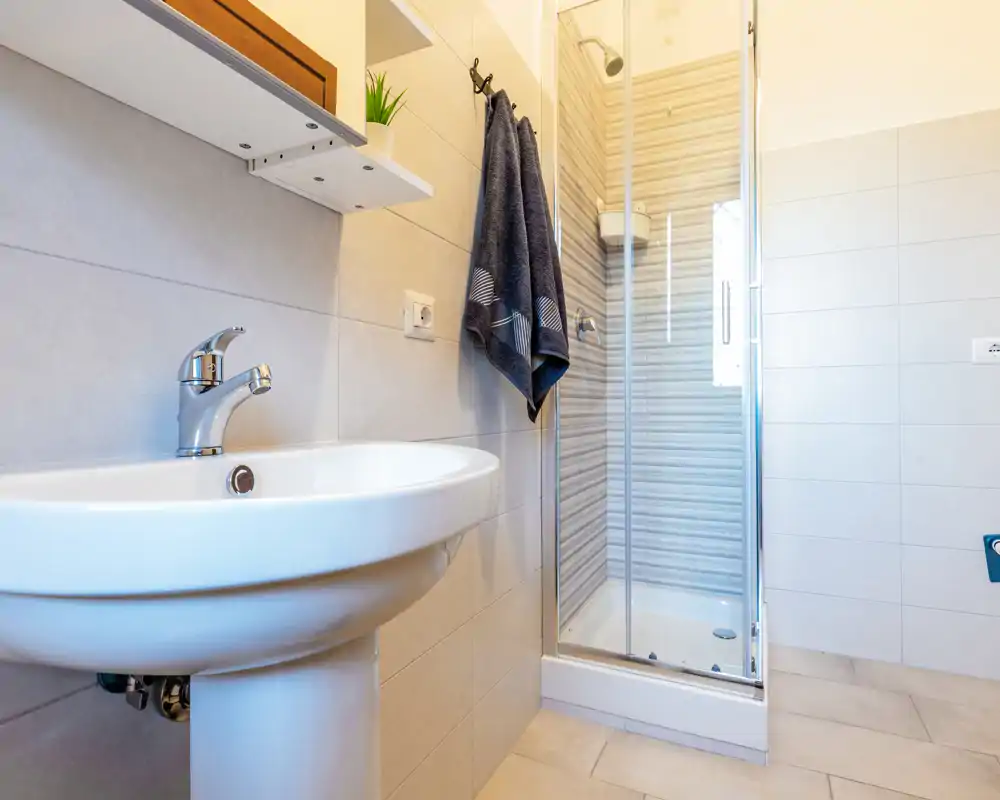 Bagno moderno e pulito con lavabo a colonna e doccia con box in vetro; piastrelle neutre e luce brillante creano un ambiente fresco e accogliente.