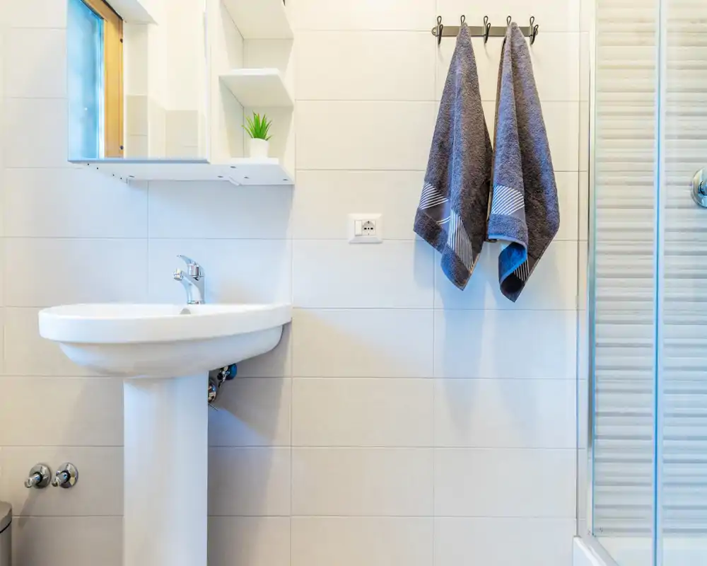 Angolo bagno pulito e moderno con lavabo a colonna, specchiera con mensole e asciugamani appesi accanto alla doccia in vetro.