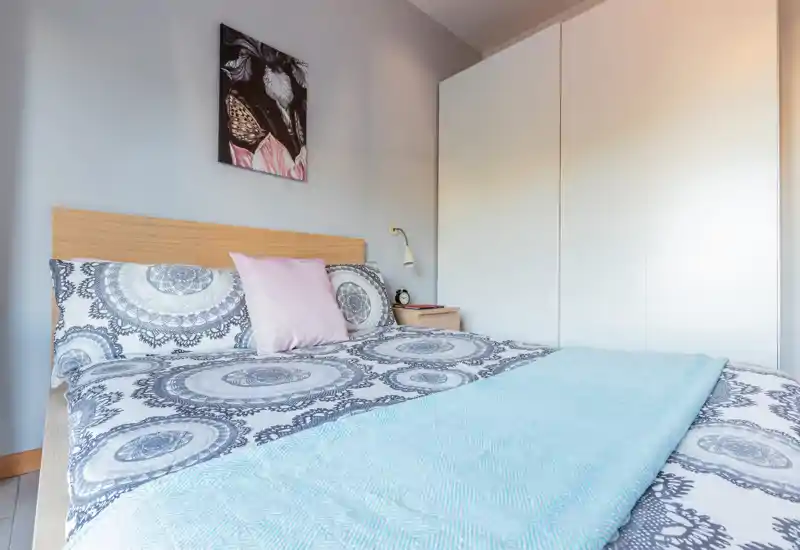 Dormitorio luminoso y ordenado con cama doble estampada, toques en tonos pastel y un armario blanco alto. La habitación se muestra acogedora y lista para un anuncio de alquiler.