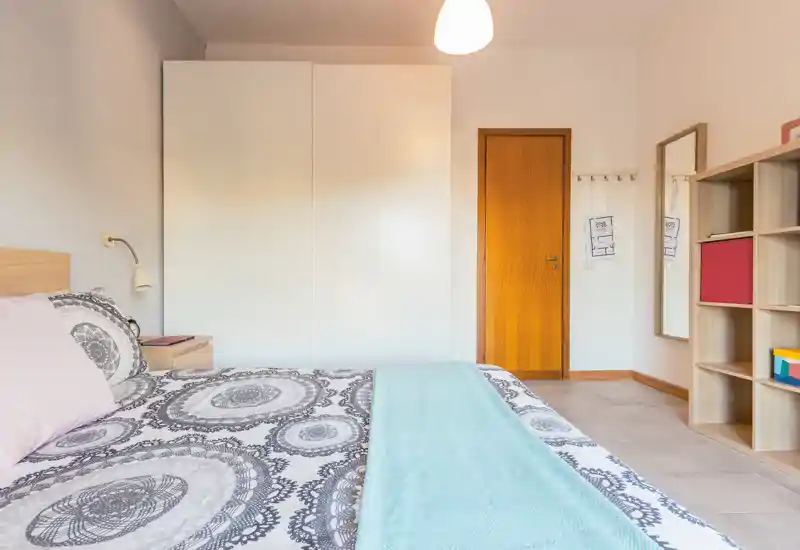 Dormitorio luminoso y ordenado con cama doble estampada, mesita de noche y armario — tonos neutros y una manta turquesa que aportan calidez.