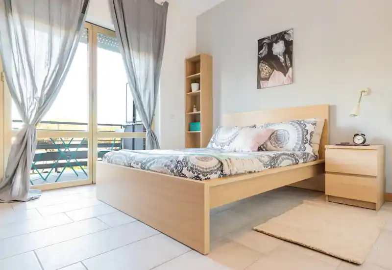 Dormitorio moderno y luminoso con cama doble de madera, ropa de cama con estampado y luz natural desde el balcón. Mobiliario minimalista y textiles suaves que crean un ambiente cálido y relajante.