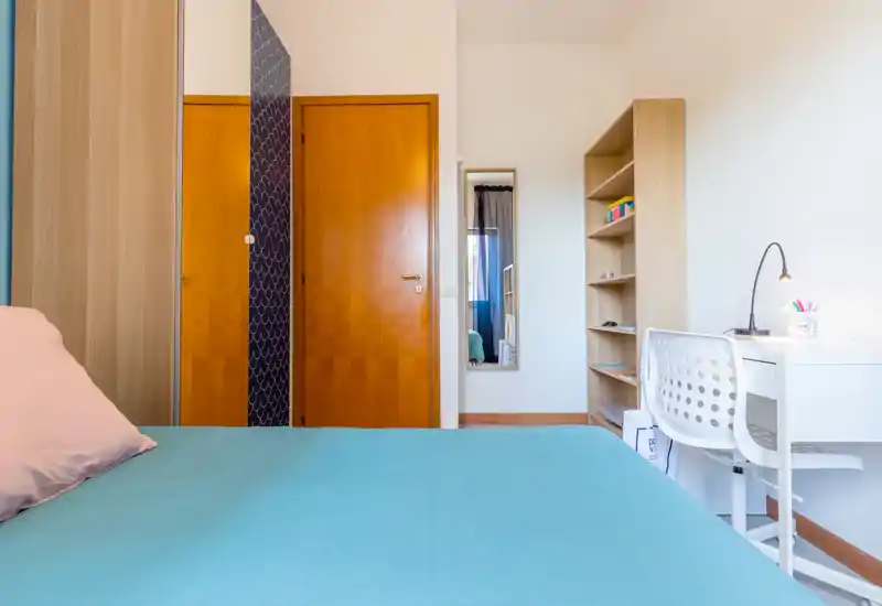 Bright, tidy bedroom with a teal bedspread, bedside pillow and a compact white desk with shelving — ideal for a student or single occupant.