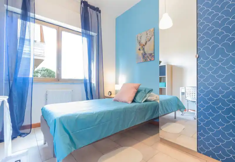 Bright single bedroom with a turquoise bedspread, soft pillow accents and a large window letting in abundant natural light — ideal for a comfortable stay.