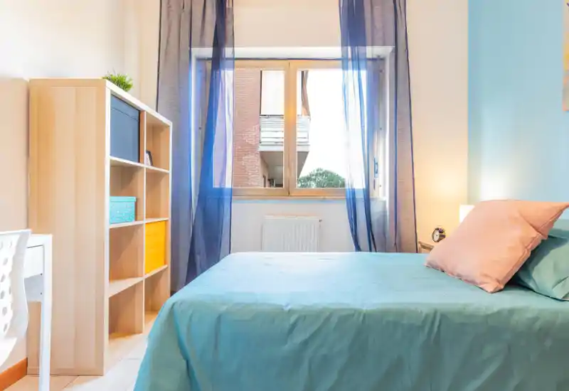 Bright, well-staged bedroom with a neatly made double bed, soft blue linens, bedside lamp and a shelving unit beside a large window letting in natural light.