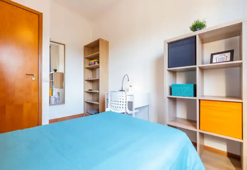 Bright, tidy bedroom with a blue-covered bed, shelving units and a small desk — neat, well-lit space ideal for a single occupant or student.