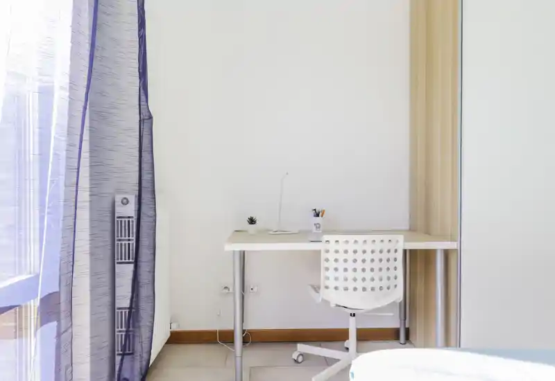 Bright minimal corner of a bedroom with a simple white desk, perforated swivel chair and sheer curtains letting in natural light — ideal as a compact workspace.