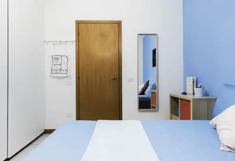 Bright modern bedroom with a large blue bed in the foreground, a wooden door, full-height wardrobe and a mirror reflecting the room. Clean, airy styling with colorful cushions and a small bedside shelving unit.
