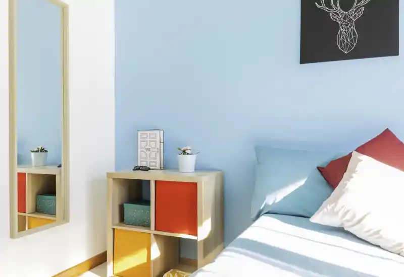 Bright, modern bedroom corner with turquoise wall, colorful cushions and a small cube shelf next to the bed — fresh, well-lit and stylish.