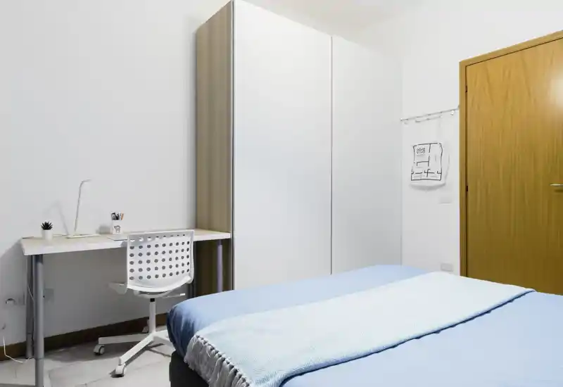 Bright, minimal bedroom with a large bed dressed in light blue linens, a simple desk and wardrobe — clean, airy and well-lit space ideal for rest and study.