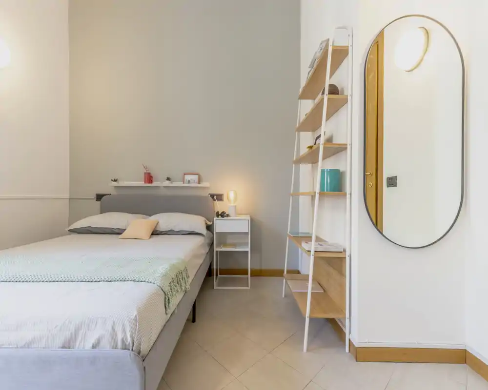 Bright, modern bedroom with a neatly made double bed, bedside table and ladder-style shelving; neutral tones and soft textiles create a calm, welcoming atmosphere.