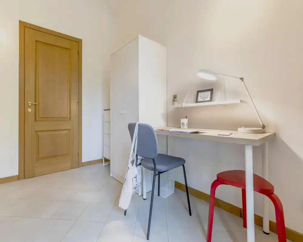 Small, tidy bedroom corner with a work desk, chair and wardrobe. Bright neutral tones and a focused desk lamp create a functional study area within the sleeping space.