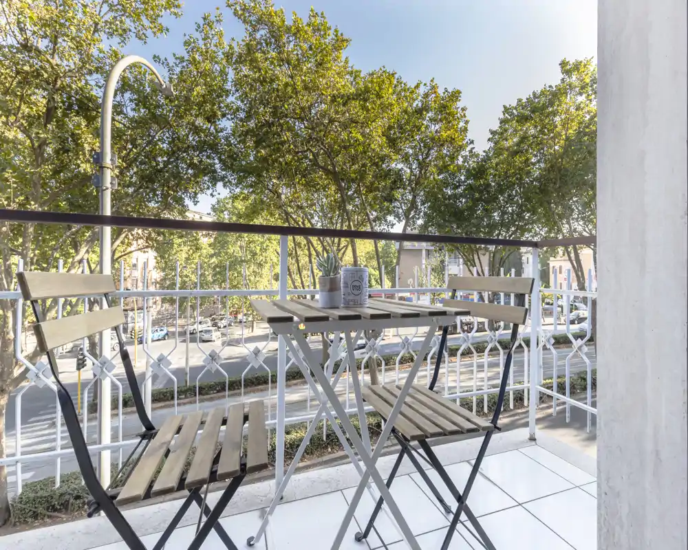 Sunny balcony with a small bistro table and two folding chairs, overlooking a tree-lined street—ideal for morning coffee or relaxing outdoors.