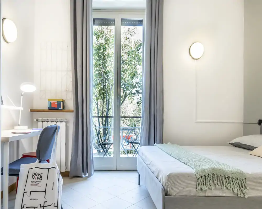 Bright, modern bedroom with a comfortable double bed, soft throw and direct access to a small balcony; a desk and chair create a functional workspace by the window.
