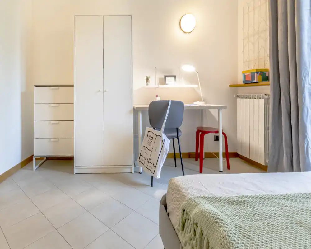 Bright, softly styled bedroom corner with a visible bed corner, wardrobe and a tidy desk with lamp — ideal for a single occupant or student.