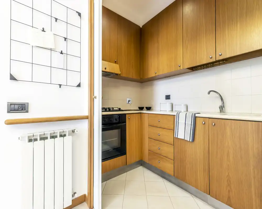 Compact L-shaped kitchen with warm wooden cabinets, integrated oven and gas hob, tiled backsplash and a stainless sink — bright and functional space ideal for city apartments.