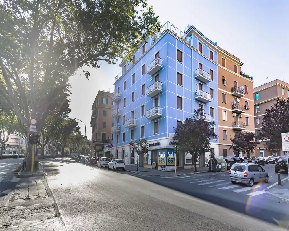 Exterior street view of a corner building with a blue façade, small balconies and ground-floor shops, tree-lined avenue and parked cars providing an urban neighborhood context.
