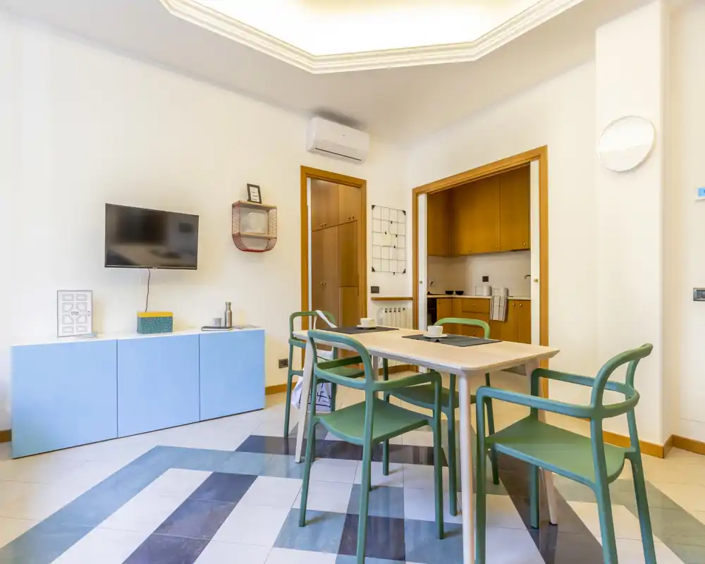 Bright, modern living/dining area with a wooden table, green chairs and a wall-mounted TV; open access to a compact kitchen.
