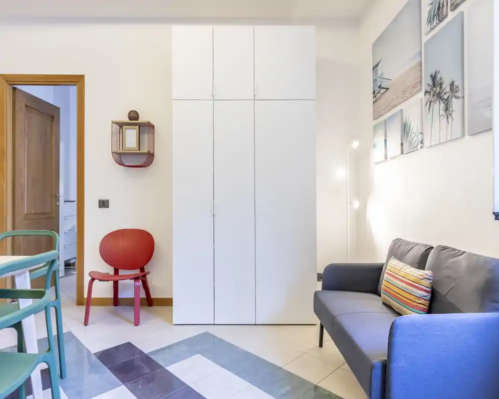 Bright, modern living area with a compact sofa, colorful cushion and framed coastal artwork; tasteful storage and seating create a cozy, well-styled space.