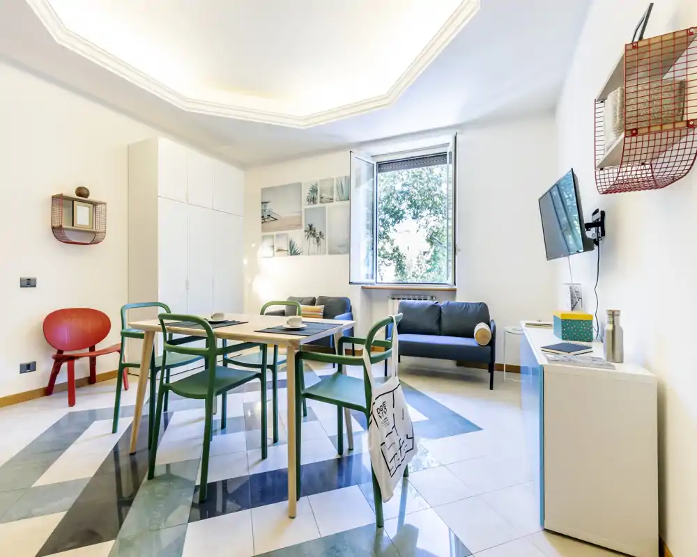 Bright, modern living/dining area with a central dining table, green chairs and a comfortable sofa by an open window letting in natural light.