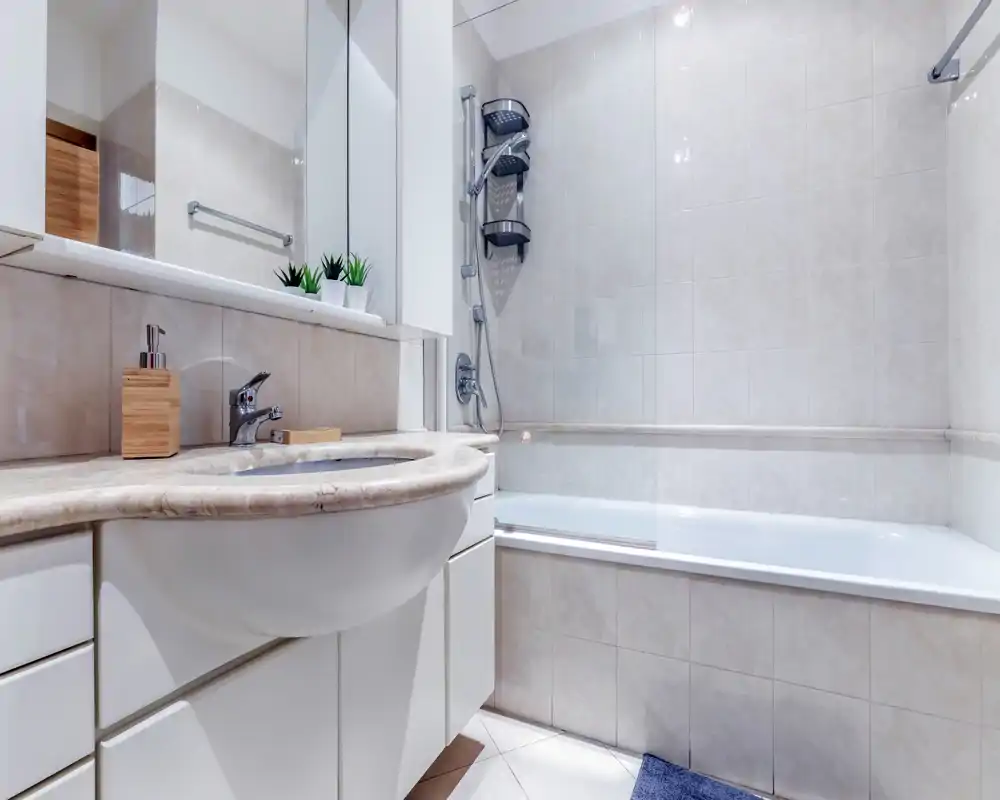 Clean, modern bathroom with a built-in bathtub, wall-mounted showerhead, sink with marble countertop and mirrored cabinet; neutral tiled walls and bright lighting.
