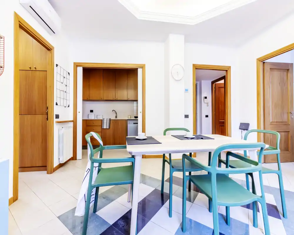 Bright, modern dining area with a simple wooden table and green chairs placed on a patterned tile floor; the open doorway reveals a compact kitchen, creating an open-plan living feel.