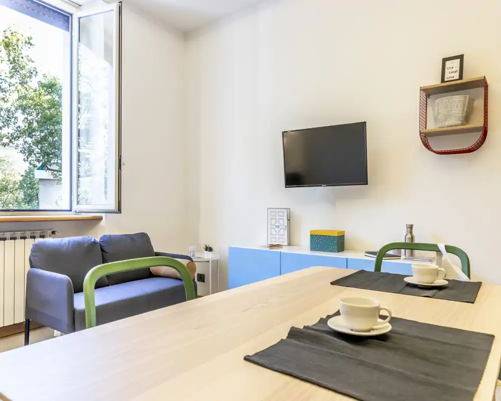 Bright, modern living area with a small sofa, wall-mounted TV and a dining/work table by an open window—clean styling and natural light create a welcoming space.
