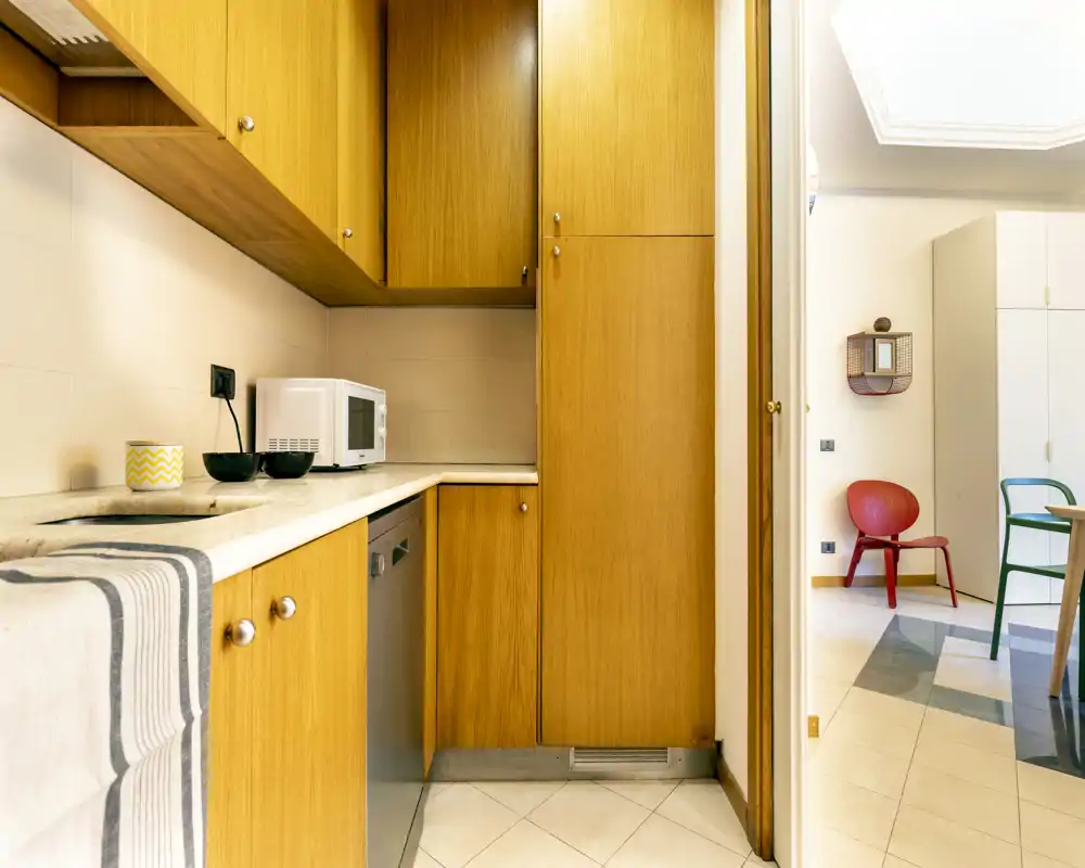 Compact kitchen with warm wooden cabinetry, countertop microwave and integrated dishwasher; bright, tidy layout connecting to an adjacent dining or living area.