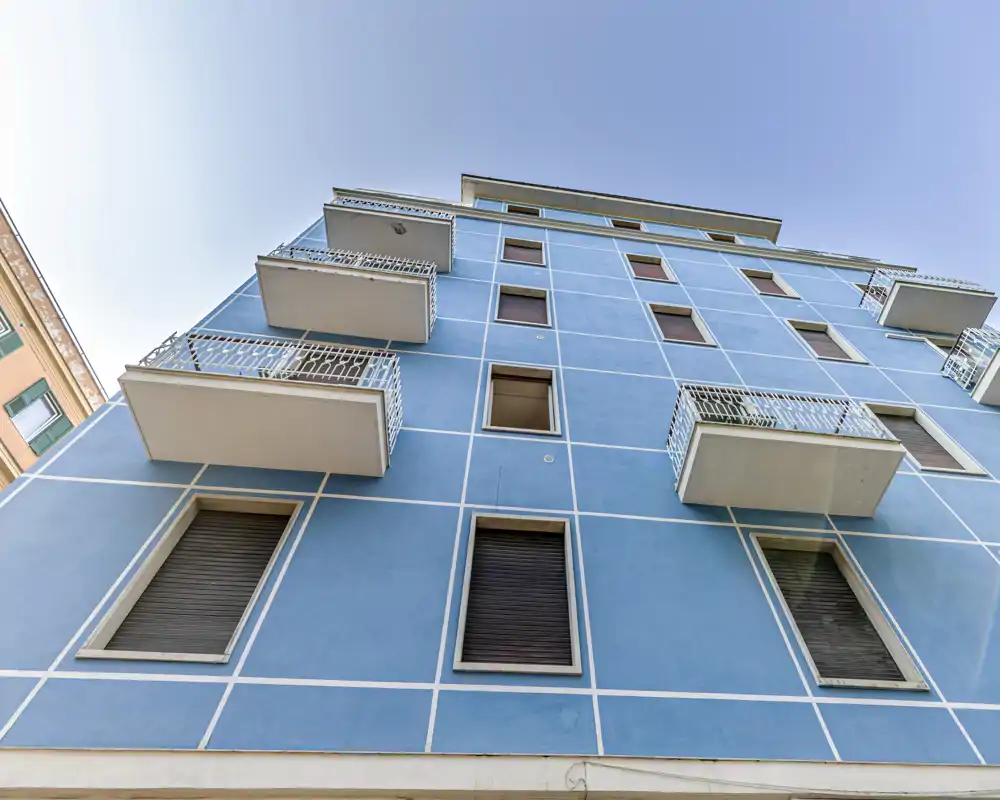 Blue apartment building façade photographed from below, showing multiple balconies and closed windows against a clear sky.