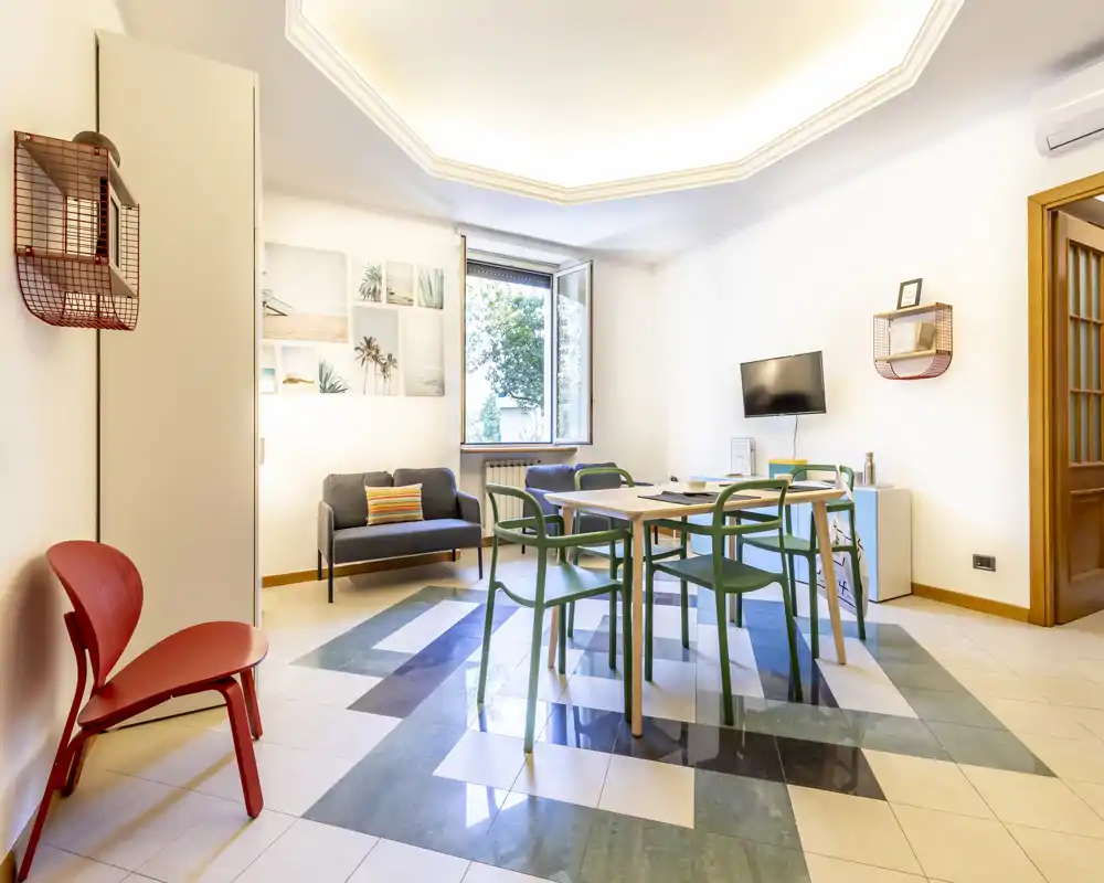 Bright, modern living/dining area with a central table and green chairs, a small sofa by the window, decorative wall art and polished patterned flooring.