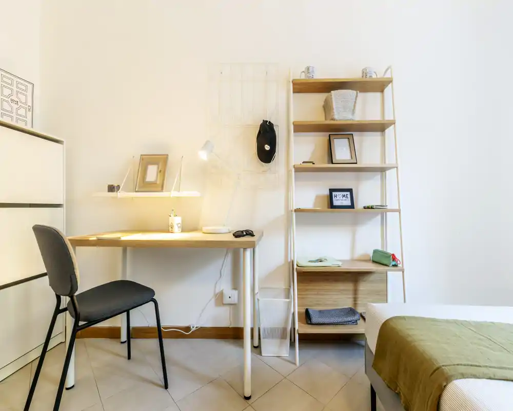 Bright, minimal bedroom corner with a visible bed edge, a tidy desk and shelving — ideal for a single occupant or student. The warm desk lamp and soft textiles create an inviting, well-lit workspace beside the sleeping area.