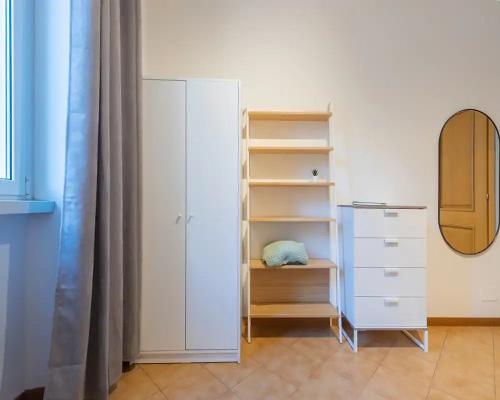 Minimal bedroom corner with a wardrobe, open shelving and a chest of drawers beside a window; simple, bright and neatly arranged storage area.