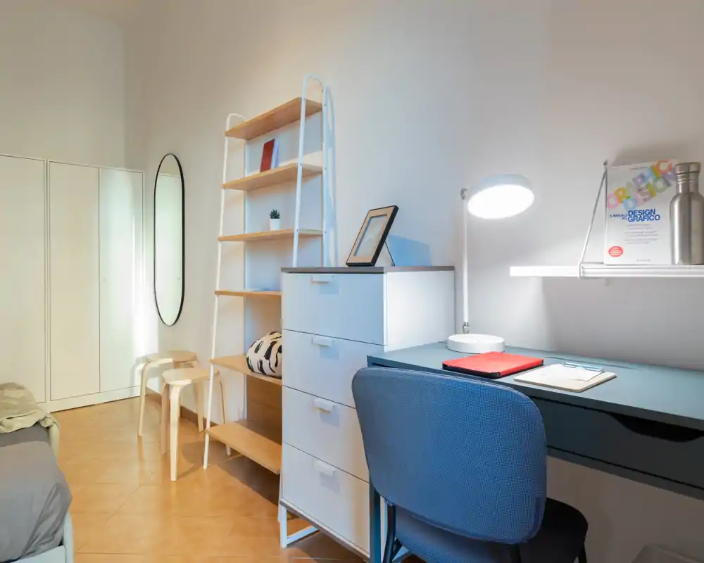 Bright, tidy bedroom corner with a study desk: a blue desk and chair, shelving and drawers, plus a partially visible bed at the left. The image highlights a functional workspace inside the sleeping area.