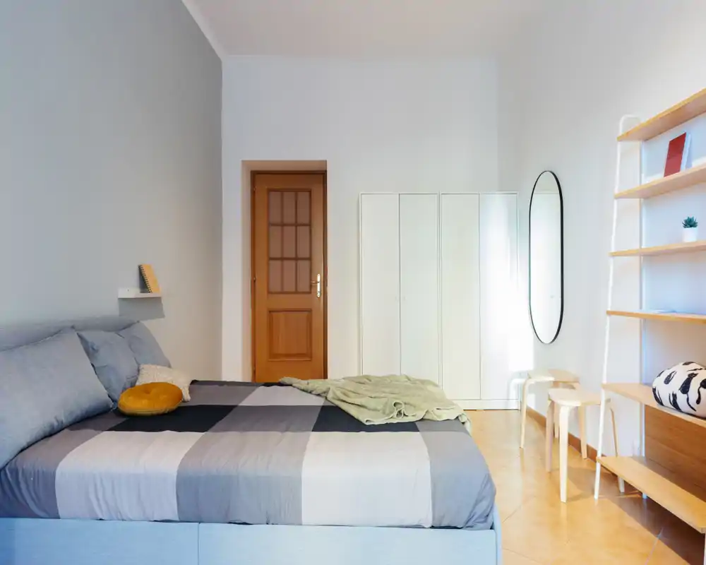 Bright, modern bedroom with a large double bed dressed in neutral linens, a white wardrobe, wall mirror and minimalist shelving — ideal for a restful stay.